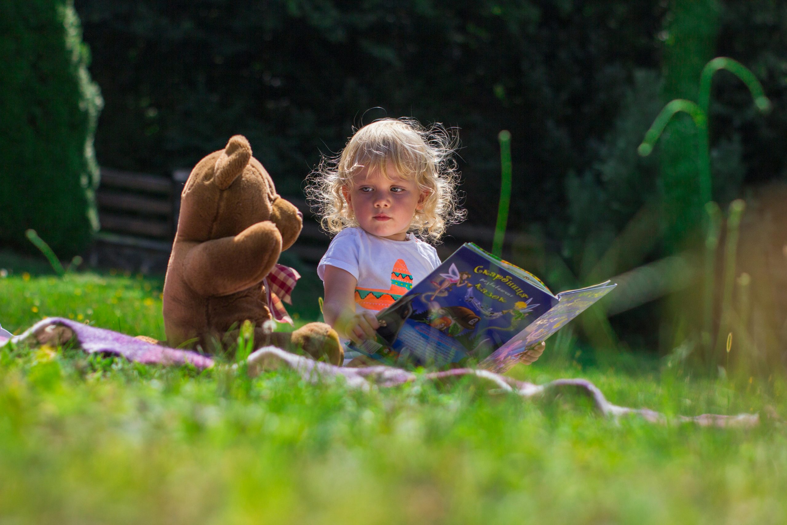 young girl holding a book while sitting outdoors with a teddy bear next to her